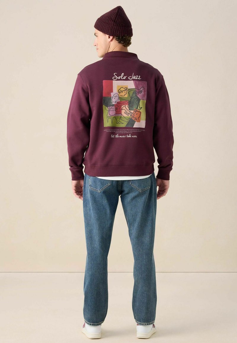 Bordeauxfarbenes Sweatshirt mit grafischem Druck auf dem Rücken, das abstrakte Figuren zeigt. Kombiniert mit blauen Jeans und weißen Turnschuhen.