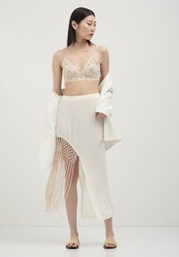 Bralette en dentelle beige clair associé à une jupe blanche transparente avec des franges sur les côtés et des détails en filet, porté avec une couche extérieure blanche.