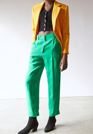 Person trägt ein gelbes Sakko, ein schwarzes, geknöpftes, kurzes Top, leuchtend grüne, hoch taillierte Hosen und schwarze Stiefeletten, steht auf einem hellen Boden.