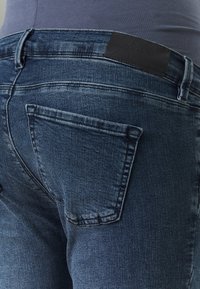 Jeans de mezclilla azul oscuro con un bolsillo trasero, ligero desgaste y una textura agradable. Presenta un logo de parche de cuero negro en la cintura.