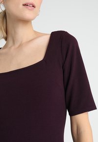 Kortärmad topp i mörklila, med fyrkantig halsringning och texturerad tyg, i en figurnära passform och minimala sömdetaljer.