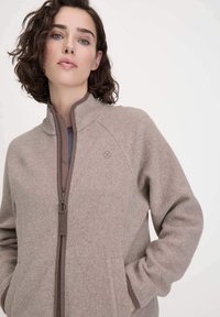 Hellbraune Fleecejacke mit hohem Kragen, Reißverschluss und seitlichen Taschen. Sieht mit kontrastierenden braunen Abschlüssen und einem dezenten Logo auf der Brust aus.