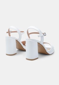 Valkoiset korkokenkä-sandaalit, joissa on paksu lohkokorko, avoin varvas ja ohuet nilkkahihnat. Kehyksetön synteettinen materiaali, jossa on beige pohjallinen.