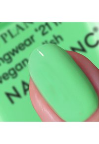 Ilar grön nagellack med en glänsande finish på en fingertopp, med en suddig bakgrund som visar produktetiketter och detaljer.