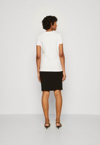 Top blanco de manga corta combinado con una falda negra hasta la rodilla, llevado con sandalias negras, mostrado desde la parte posterior contra un fondo liso.