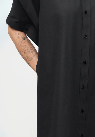 Τατουάζ στον βραχίονα με λεπτομερείς ανθρώπινες φιγούρες κλασικού στυλ σε άτομο που φοράει μαύρο, χαλαρό πουκάμισο με κουμπιά και μανίκια γυρισμένα.