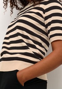 Person iført en beige og sort vandretstribet T-shirt med kortærmer og den ene hånd i lommen på sorte bukser mod en hvid baggrund.