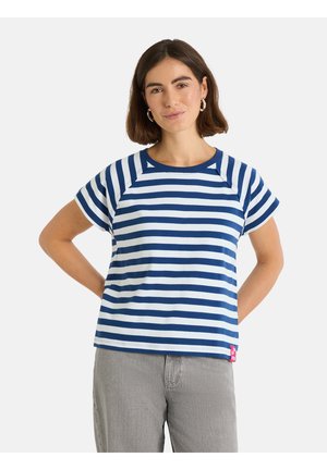 Frau mit kurzen braunen Haaren, die Creolen trägt, ein blau-weiß gestreiftes Kurzarmshirt und graue Hosen, steht mit den Händen hinter dem Rücken.