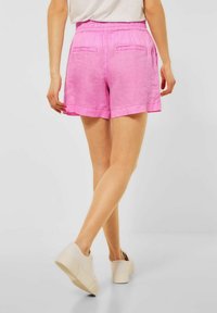 Kvinna klädd i ljusrosa shorts och vita sneakers, som står med ett ben korsat bakom det andra mot en neutral bakgrund.