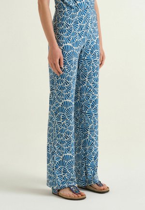 Donna che indossa pantaloni ampi con fantasia blu e bianca e sandali colorati con perline, in piedi davanti a uno sfondo semplice.