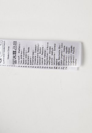 Štítek vyrobený z bílé tkaniny, který obsahuje pokyny k péči, informace o materiálu a symboly praní vytištěné v několika jazycích.