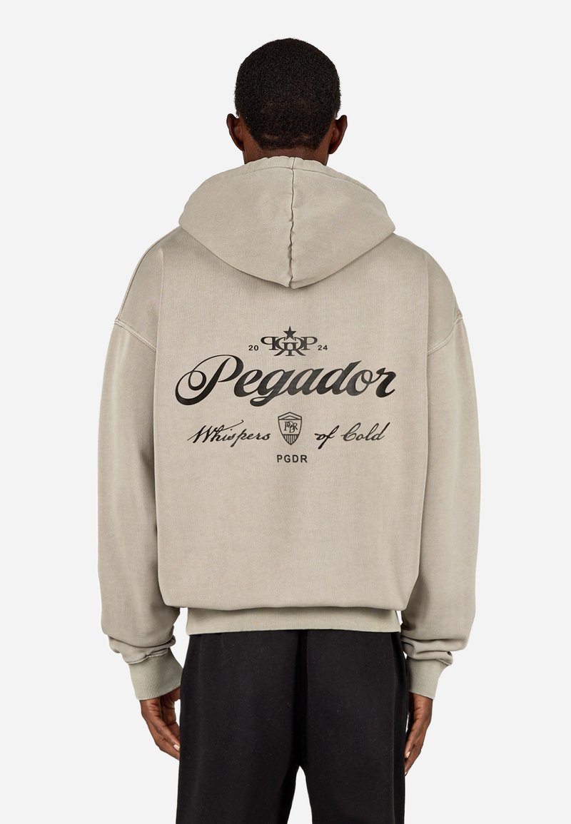 Beige oversize kapuce ar pietvērējiem un teksturētu apdari. Aizmugurē ar melnu drukātu logotipu un tekstu.