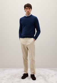 Maglione blu navy, pantaloni beige e scarpe marrone scuro. Il maglione presenta un collo rotondo; i pantaloni hanno una vestibilità su misura e una texture liscia.