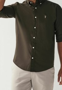 Chemise boutonnée vert foncé avec col, dotée de boutons blancs et d'un petit logo brodé sur la poitrine. Le tissu semble lisse et légèrement brillant.