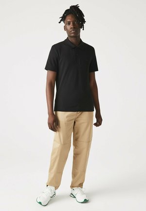 Camisa polo negra con un logo sutil, combinada con pantalones beige claros con paneles. El calzado es blanco con acentos verdes.