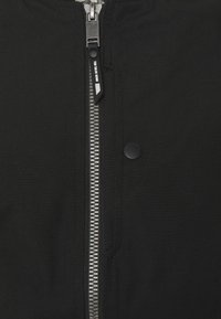 Černá bunda s výrazným stříbrným zipem, klapkou na zacvaknutí a hladkým povrchem. Vyznačuje se minimalistickým designem bez dalších vzorů.