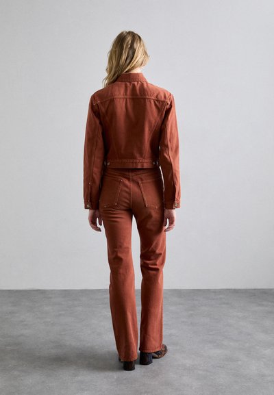 Jachetă din denim de culoare ruginită și pantaloni asortați, cu o siluetă ajustată, având buzunare la spate și accente cu nasturi, plasate pe un fundal neutru.