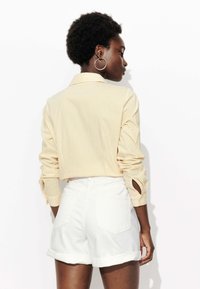 Chemise rayée jaune et blanche avec des manches retroussées, accompagnée de shorts en denim blanc. La chemise a un col et des détails de boutons.