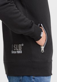Schwarzer Hoodie mit einer Reißverschlusstasche, ausgestattet mit einem grauen Logo. Hergestellt aus weichem Stoff mit gerippten Bündchen und Saum, präsentiert ein lässiges Design.
