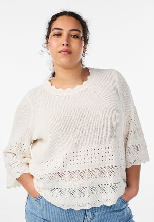 Donna con capelli ricci scuri che indossa un maglione bianco lavorato a maglia con motivi in pizzo e jeans blu, in piedi con le mani in tasca davanti a uno sfondo bianco.