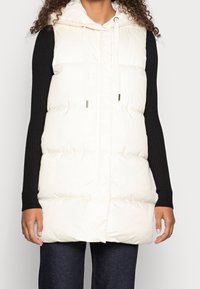 Wattierte ärmellose Weste in Creme mit Kapuze, ausgestattet mit gerippten schwarzen Ärmeln und Kordelzugakzenten. Getragen über einem dunklen Oberteil und Jeans.