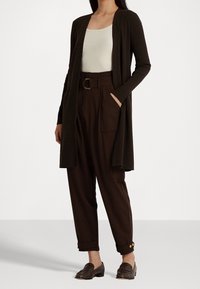 Lungo cardigan marrone sopra una maglia beige chiara, abbinato a pantaloni scuri a vita alta in marrone con grandi tasche e polsini con bottoni. Mocassini marroni.