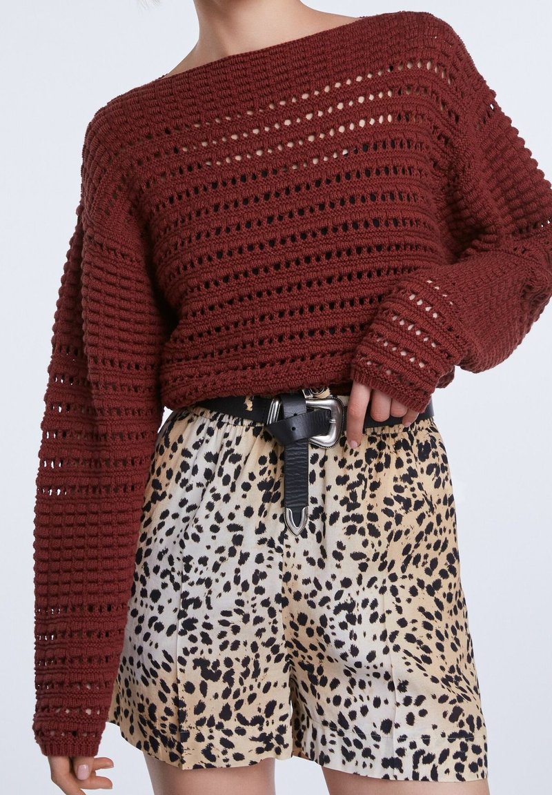 Personne portant un pull en crochet bordeaux rentré dans un short taille haute beige à pois noirs avec une ceinture noire et une boucle argentée.