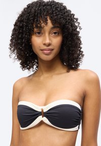 Νεαρή γυναίκα με σγουρά μαλλιά που φοράει μαύρο μπικίνι χωρίς τιράντες με λευκή επένδυση, κοιτώντας την κάμερα με ουδέτερη έκφραση.