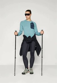 Maglietta a maniche lunghe color teal con una tasca blu scuro, leggins neri e scarpe da trekking grigie. La persona tiene dei bastoni da trekking ed indossa occhiali da sole scuri.