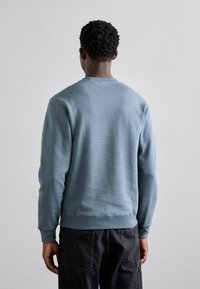 Hombre con cabello rizado corto, vistiendo un suéter azul claro y jeans negros, de pie frente a una pared lisa de color gris claro.