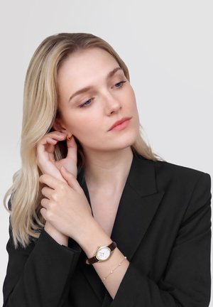 Plavuša u crnom sakou prilagođava zlatnu naušnicu u obliku obruča, pokazujući ručni sat s smeđim kožnim remenom i nježnu narukvicu.