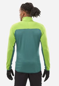 Person with dark braided hair and ponytail wears green and teal long-sleeve top and black gloves, shown from the back on a white background.