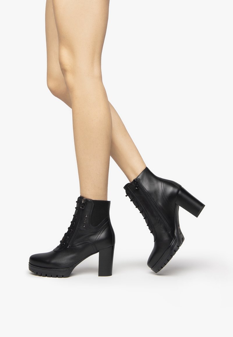 Jambes portant des bottines en cuir noir à lacets, avec des fermetures éclair latérales et des talons hauts épais, sur fond blanc.
