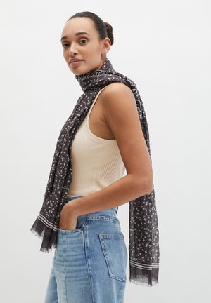 Vrouw met haar in een knot, gekleed in een mouwloze beige top, blauwe spijkerbroek en een donkere sjaal met bloemenpatroon om haar nek, tegen een effen achtergrond.