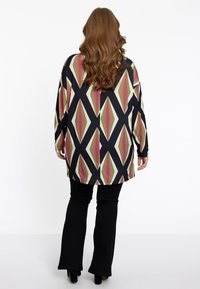 Schwarze Tunika mit langenÄrmeln, versehen mit einem geometrischen Diamantenmuster in Grün und Pink. Kombiniert mit figurschmeichelnden schwarzen Schlaghosen und High Heels.