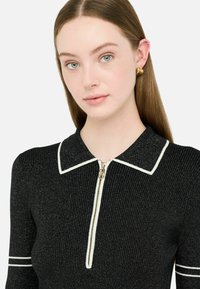Čierny žebrovaný top na zips s bielym golierom a akcentmi. Obsahuje kovový zips s okrúhlym ťahákom. Mäkká, textúrovaná látka.