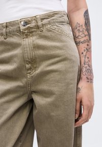 Κοντινό πλάνο μπεζ παντελονιού με λεπτομέρεια κουμπιού και τατουάζ στο χέρι που στηρίζεται δίπλα στο ισχίο, φορώντας λευκό τοπ.