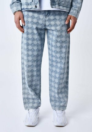Pantalon en denim bleu clair avec un motif en diamants, coupe ample et coutures visibles. Associé à des chaussures de sport blanches.