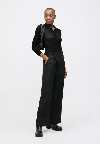 Crna pletivna polo majica s ovratnikom i gumbima, uparen s širokim crnim hlačama s kosim prugama i debelim crnim cipelama.