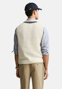 Mies, joka wear navy-lippisme, vaalea neuleliivi, siniraitainen paita, jonka hihat on kääritty, ja beigelhousut, näkyy selkäpuolelta.