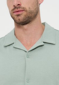 Άνδρας με ελαφριά γενειάδα που φοράει πουκάμισο σε παστέλ πράσινο χρώμα με γιακά και δύο ορατά κουμπιά σε λευκό φόντο.