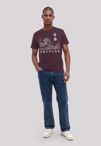 Burgundy cotton t-shirt featuring white Japanese text and mountain graphic, paired with blue jeans and white sneakers.
