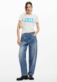 T-shirt bianca con grafica blu "LOVE DSGL", abbinata a jeans in denim blu larghi e strappati e stivali platform neri.