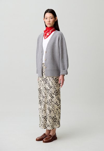 Cárdigan gris de punto con botones dorados sobre una blusa blanca, falda larga estampada en crema y negro, y mocasines marrones. Pañuelo rojo atado al cuello.