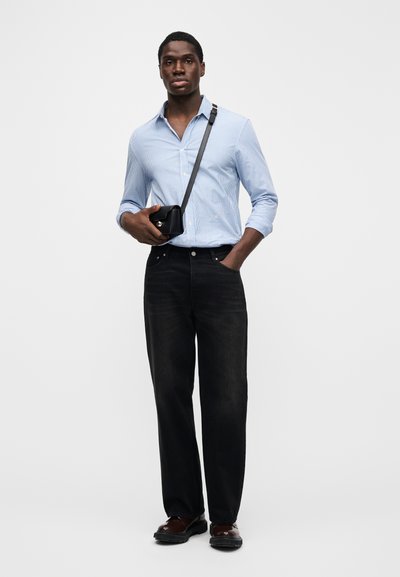 Blåt og hvidt stribet skjorte med knapper, sorte jeans, sort skuldertaske, brune sko, stående positur mod en neutral baggrund.