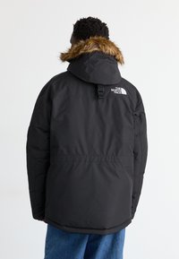 Osoba w czarnej ocieplanej kurtce z futrzanym kapturem i logo The North Face na górnej części pleców, w zestawieniu z niebieskimi dżinsami.