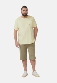 Hellgelbes Baumwoll-T-Shirt mit kurzen Ärmeln und einer Brusttasche, kombiniert mit beigen Baumwollshorts und weißen Sneakers auf einem schlichten Hintergrund.