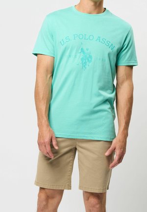 Mann trägt ein hellblaues U.S. Polo Assn. T-Shirt mit Logo und beige knielange Shorts, steht vor einem einfarbigen Hintergrund.