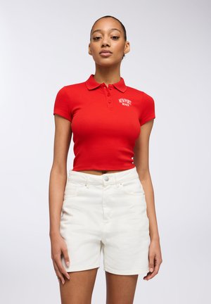 Polo shirt corto rosso con maniche corte, presenta "NEWPORT BEACH" ricamato in bianco. Abbinato a pantaloni corti in denim bianchi.