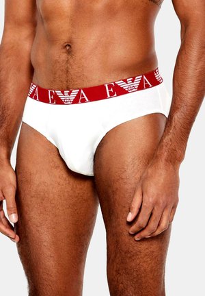 Białe majtki typu briefs z czerwonym elastycznym paskiem w talii ozdobionym powtarzającym się wzorem logo. Materiał wydaje się gładki, a krój dopasowany.